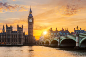 A scenic view of Big Ben and the Houses of Parliament in London, England, at sunset representing a perfect London weekend itinerary. The River Thames flows in the foreground with a bridge crossing it.