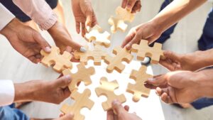 Group of hands connecting wooden puzzle pieces in a circle, symbolizing teamwork and collaboration.
