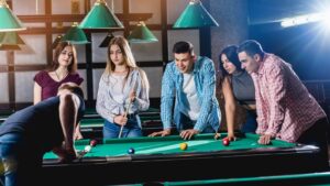 A group of young adults playing pool, with one person taking a shot while others watch, under green-shaded lights in a lively room.