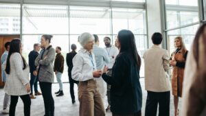 A large group of business professionals are networking and chatting in a bright, modern venue with large windows. Text: "What Is Corporate Hospitality." This image shows a typical London corporate event for businesses.