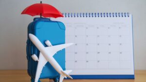 A small blue suitcase with a red umbrella on top is next to a calendar, with a white toy airplane resting against the suitcase.