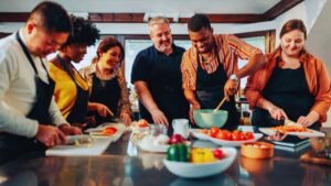 Diverse group in aprons enjoys team cooking class: chopping veggies like tomatoes & peppers, stirring bowls, collaborating in bright kitchen with fresh ingredients.