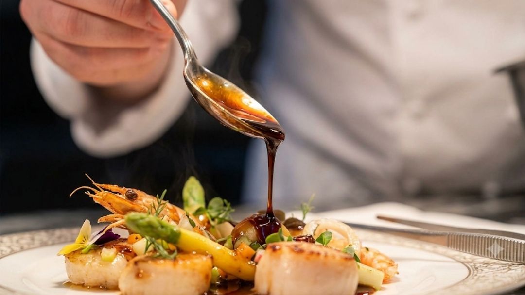 Galvin La Chapelle French private dining for the City: a chef spoons glossy sauce over a plated dish of seared scallops, prawns, and vegetables in close-up.